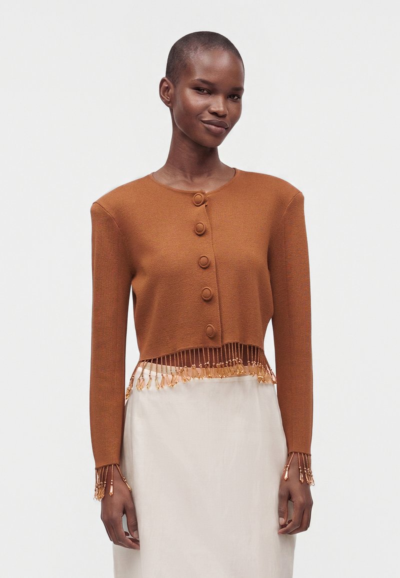 Woman wearing a brown buttoned cropped sweater with beaded fringe on hem and cuffs, paired with a light beige skirt against plain background.