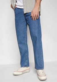 Man med blå raka jeans, vita sneakers med tjocka sulor, en vit t-shirt och en beige rutig skjorta med händerna vid sidorna.