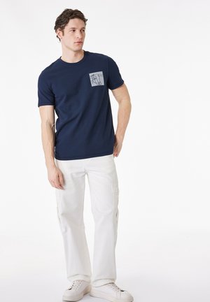 Mężczyzna ubrany w granatowy t-shirt z kieszenią z wzorem konia, białe spodnie typu cargo i białe trampki, stoi na białym tle.