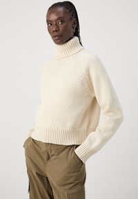 Cremefarvet turtleneck sweater med ribbet kant og manchetter; kraftig strikket tekstur; afslappet pasform; parret med olivengrønne bukser.