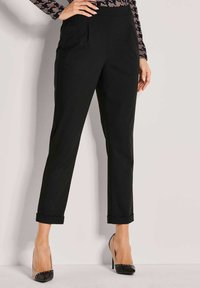 Vrouw in zwarte op maat gemaakte cropped pants met manchetvoeten en zwarte hoge hakken met naakte zijkanten, staande tegen een effen muur.