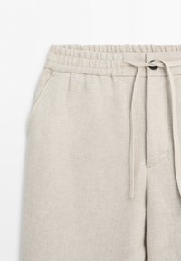 Beige linen pants with elastic waistband, drawstring, button, and side pocket, shown from the waist to mid-thigh on a white background.