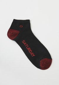 Schwarze Socke mit rotem Zeh und Ferse, versehen mit dem Schriftzug "SAMSTAG" in rotem Text an der Seite. Hergestellt aus weichem, strukturiertem Stoff.