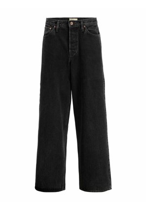 Personne portant un t-shirt blanc uni à manches courtes, un jean noir ample et des baskets beige et blanches, debout sur un fond blanc.