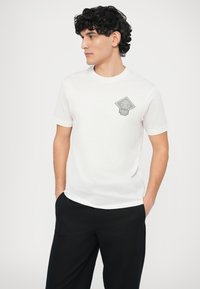 Biały bawełniany t-shirt z okrągłym dekoltem, z szarą naszywką w kształcie diamentu na lewej piersi. Krótkie rękawy, gładka faktura.