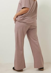 Lös passform, ljus lila loungeklädesset som består av en kortärmad topp och vida byxor i mjukt, strukturerat tyg. Svart fotbeklädnad synlig.