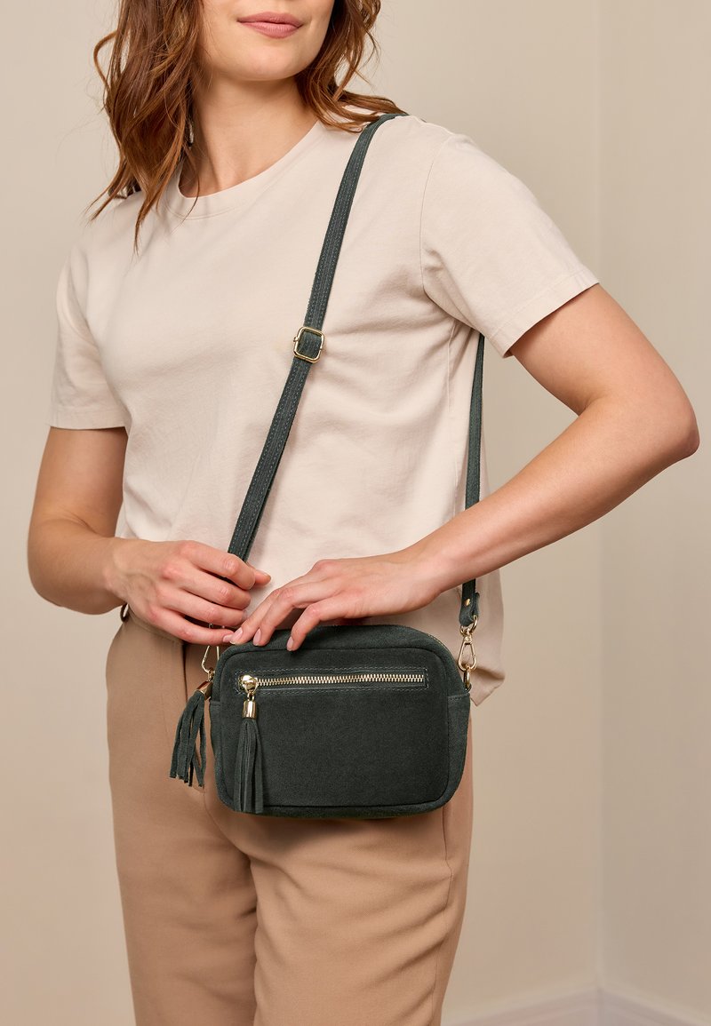 Zelená semišová kabelka přes rameno s kapesním zipem, ozdobnými třásněmi a odnímatelným popruhem. Kabelka má obdélníkový tvar a zlaté kovové prvky.