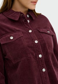 Burgundská kordová košile s button-up přední částí, dvěma kapsami na hrudi a límečkovým výstřihem, s texturovanými vertikálními pruhy a stříbrnými knoflíky.