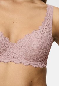 Lace bh i mauve med bølgede kanter, prydet med et blomstermotiv og en central knappedetalje. Blød tekstur og dekorative stropper.