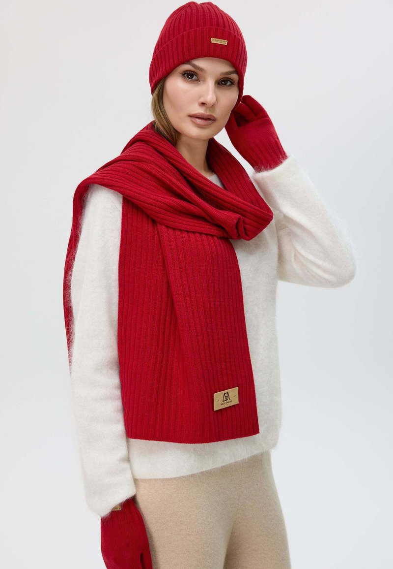 Red ribbed knit scarf with matching beanie and gloves, featuring a textured finish and a small logo tag. Worn over a white sweater.