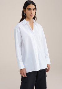 Camicia bianca a maniche lunghe con colletto classico, caratterizzata da una vestibilità rilassata e una consistenza fresca, abbinata a pantaloni neri.