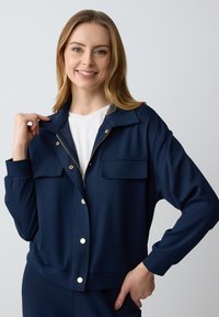 Giacca con zip navy realizzata in tessuto liscio, caratterizzata da bottoni a pressione in tonalità oro e due tasche sul petto, con una vestibilità rilassata e maniche lunghe.