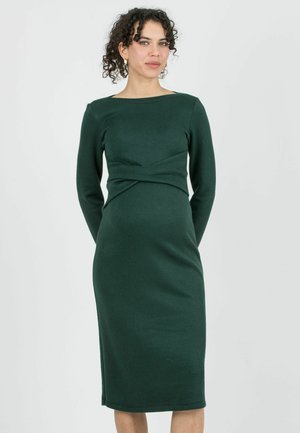 Vrouw met krullend haar draagt een aansluitende, donkergroene midi-jurk met lange mouwen, staand tegen een effen lichte achtergrond.