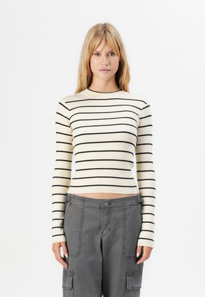 Woman with long blonde hair wearing a cream ribbed sweater with black stripes and gray cargo pants, standing against a white background.