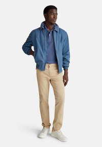 Niebieska bombowiec z kołnierzem, nałożona na granatowy polo, w zestawie z beżowymi spodniami i białymi trampkami z żółtymi akcentami.