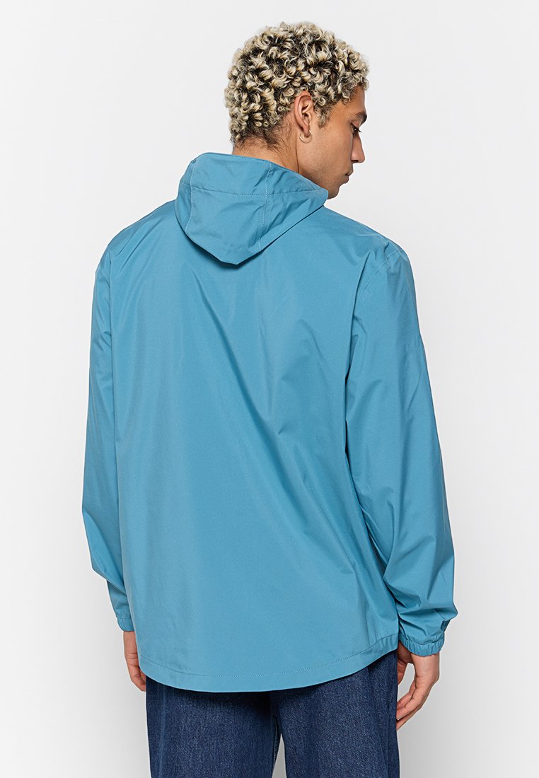 Person mit lockigem blonden Haar, der eine blaue Kapuzenjacke trägt und von hinten zur Kamera schaut, kombiniert mit dunklen blauen Hosen vor einem schlichten weißen Hintergrund.