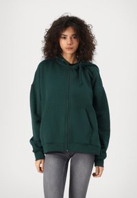 Zelena zip-up majica od mekog materijala, s kenguretorom, kapuljačom na vezanje i rebrastim rukavima. Nosio se s uskim sivim trapericama.