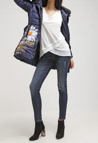 Cappotto imbottito blu navy con ricamo floreale, top bianco a strati, jeans skinny scuri e stivali neri alla caviglia con accenti metallici.