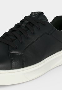 Baskets en cuir noir avec lacets noirs et semelle en caoutchouc blanche, présentant des coutures détaillées et une languette matelassée avec un logo discret.