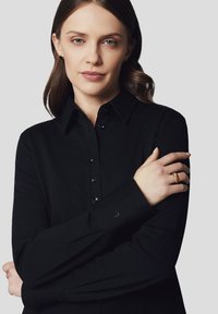 Czarna koszula z guzikami, wykonana z teksturowanej tkaniny, z długimi rękawami i spiczastym kołnierzykiem. Posiada czarne guziki oraz subtelne logo na mankiecie.