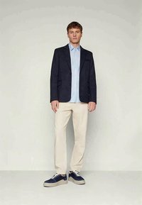 Navy blazer over light blue striped shirt, beige trousers, and navy sneakers. Features a classic fit and casual style. Smooth textures.