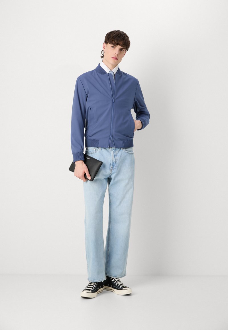 Veste zippée bleue avec des poignets côtelés, associée à un jean en denim bleu clair et des baskets noires, tenant une tablette noire à la main. Fond uni.