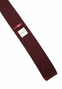 Burgundy stickad slips med en läderslinga och en vit etikett med "John Henric Sweden" broderad på vit bakgrund.