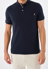 Námorně modré polo tričko vyrobené z bavlny, se zapínáním na dva knoflíky, krátkými rukávy a náprsní kapsou se červenými a bílými akcenty.