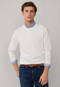 Witte gebreide trui met een ronde hals, gedragen over een blauw-wit gestreefd overhemd. Donkerblauwe spijkerbroek en een bruine leren riem maken de outfit compleet.