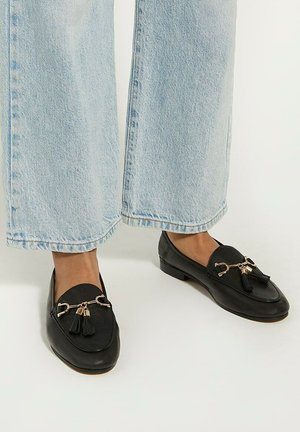 Black leather loafers with rounded toes, gold-tone hardware, and tassel accents, paired with light blue wide-leg jeans.