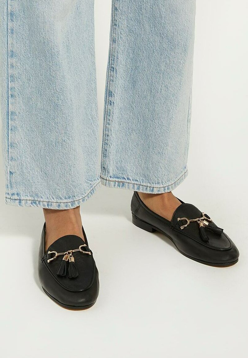 Black leather loafers with rounded toes, gold-tone hardware, and tassel accents, paired with light blue wide-leg jeans.