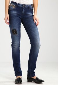 Jean en denim bleu foncé avec une coupe slim, mettant en avant des patchs décoratifs, un léger décoloré et un design classique à cinq poches. Des chaussures noires complètent la tenue.
