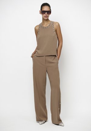 Vrouw draagt een bruin mouwloos topje en bijpassende wijde broek met metalen oogjesdetails, zonnebril en puntige schoenen van slangenleer.