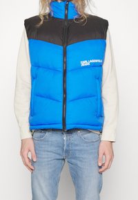 Person som bär en svart och blå vadderad väst med dragkedja fram och "KARL LAGERFELD JEANS"-logotyp, beige skjorta och ljusblå jeans.