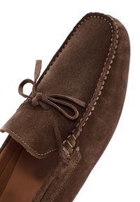 Mocasin de ante marrón con punta redondeada, acentos de costura y un detalle de lazo en la parte superior. Forro interior suave visible.
