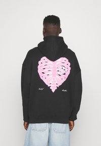 Mies joka pukeutuu mustaan huppariin, jossa on vaaleanpunainen kylkirankakuvio, ja vaaleansinisiin farkkuihin, seisoo selkä kameraan päin yksinkertaista taustaa vasten.