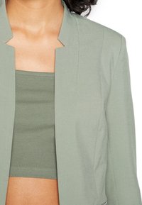 Chaqueta recortada verde claro con un escote definido y un ajuste a medida, combinada con una camiseta sin mangas de canalé a juego. Textura de tela suave.