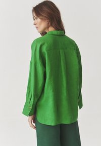Green linen shirt with long sleeves, featuring a back pleat, button cuffs, and a curved hem. Smooth texture enhances drape and fit.