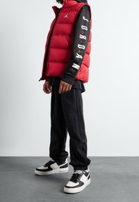 Rød puffervest med høj krave og hvidt logo, sat sammen med en sort langærmet top med hvid tekst og sorte joggers; sorte og hvide sneakers.