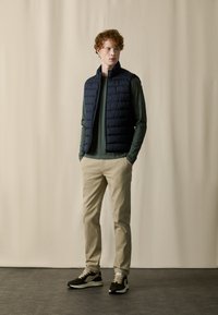 Marineblaues Puffer-Weste über einem grünen Langarmshirt, kombiniert mit beigen Hosen und schwarz-braunen Sneakers, vor einem neutralen Hintergrund.