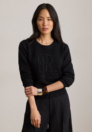 Frau mit langen dunklen Haaren, die einen schwarzen Pullover mit geprägtem Logo und schwarze Hose trägt, ergänzt durch Schildpatt- und Silberarmbänder.