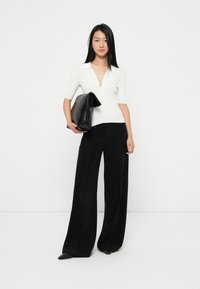 Weißes, geripptes Oberteil mit Kragen und Schnürdetail, kombiniert mit hoch taillierten schwarzen weit geschnittenen Hosen und einer zerknitterten schwarzen Clutch.