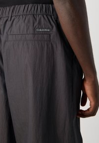 Close-up of black Calvin Klein pants with elastic waistband and back pocket on a person’s hip and arm by their side.