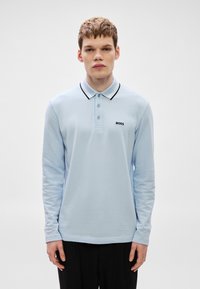 Ανοιχτό μπλε πόλο με μακριά μανίκια και μαύρο γιακά, με τρία κουμπιά και μικρό μαύρο λογότυπο στο αριστερό στήθος. Μαλακή υφή.