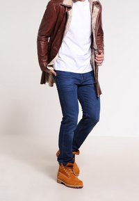 Uomo che indossa una giacca in shearling di pelle marrone, t-shirt bianca, jeans blu e stivali da lavoro beige, in piedi con una gamba incrociata sopra l'altra.
