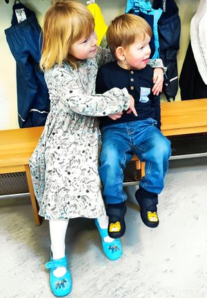 Kinder sitzen auf einer Bank, Mädchen in einem gemusterten Kleid und blauen Schuhen mit Designs, Junge in einem marineblauen Shirt und Hose, trägt schwarze Slipper mit einem Löwen.