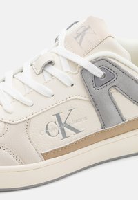 Sneakers in pelle bianca con accenti grigi e beige, dotati di linguetta imbottita, lacci e marchio "CK Calvin Klein Jeans" sui lati.