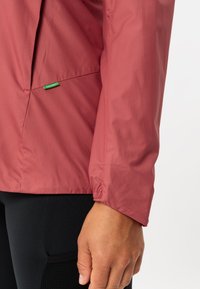 Leichte, rosafarbene Jacke mit glatter Textur, elastischen Bündchen und einem dezenten grünen Markenschild nahe der Naht. Verfügt über ein tailliertes Design.