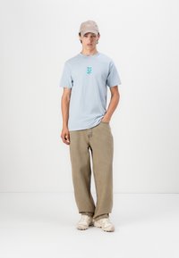 Lyseblå bomulls-T-skjorte med et turkis grafisk trykk, kombinert med beige vide bukser og lyse joggesko. Beige caps på hodet.