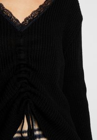 Maglione nero lavorato a maglia con scollo a V, rifinitura in pizzo e dettagli arricciati in vita con lacci regolabili. Tessuto morbido e design a coste.